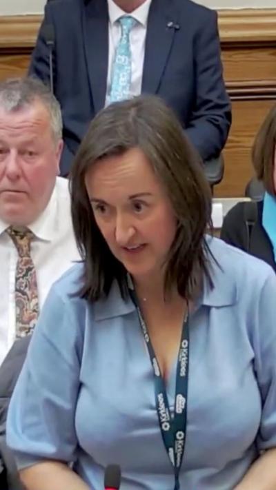 A woman with shoulder length brown hair wearing a blue shirt and lanyard, with her hand on her chest.