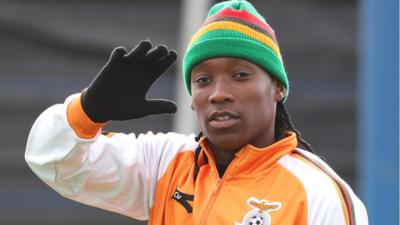 Barbra Banda in hat and Zambia tracksuit waves a gloved hand