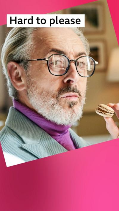 A stern looking man with grey hair holds a macaron; he wears a lilac jacket and black glasses
