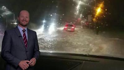 Darren Bett stands in front of dashcam footage of a flooded road.