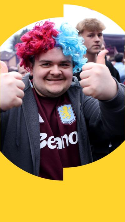Man wearing a purple and blue curly wig and an Aston Villa kit under a grey jacket. He has both thumbs up to the camera.
