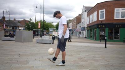 Jesse is on an almost deserted shopping street. He is wearing a white shirt and a cap and is kicking a football above his foot