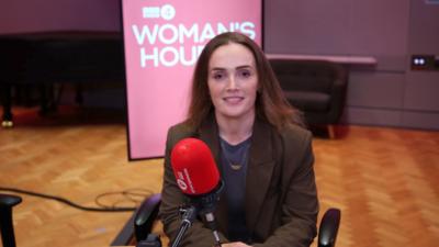 A woman with long brown hair sits in front of a red microphone.