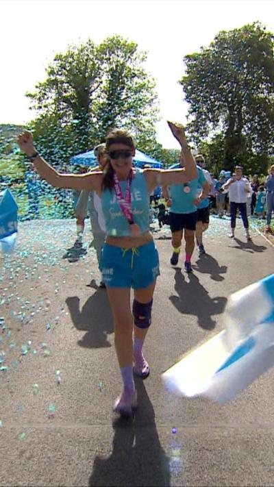 A woman is standing outside wearing a blue running outfit. She has both her arms in the air and is smiling.