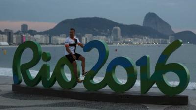 A person sits on a sign for the Rio 2016 Olympics at dawn in the Copacabana neighbourhood of Rio de Janeiro