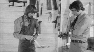 Black and white image of the subject and the reporter standing over a pot on a stove, observing the insecticide mixture brewing.  The man being interviewed is stirring the pot.  He has a bowl cut hair cut and beard and moustache and is wearing an apron..  The reporter is standing on the right hand side observicing with his arms folded and hand under his chin.