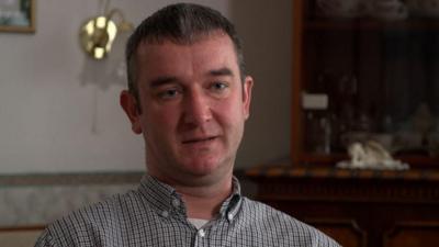 Man - Jozsef Huszti, brother of the missing sisters - wearing checked shirt and looking straight ahead while sitting in a living room