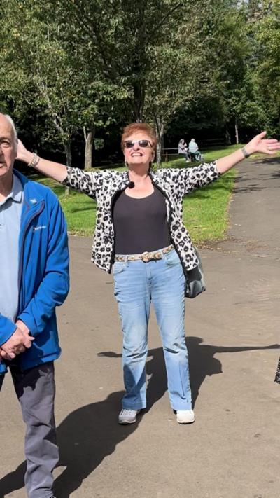 A woman stands on a sunny park path with her arms stretched wide, smiling and facing the camera. She wears sunglasses, a black top, light blue jeans, and a white-and-black patterned jacket. A man in a blue jacket and grey trousers stands beside her, slightly turned away. Behind them, trees line the path and a few people sit on benches in the distance.