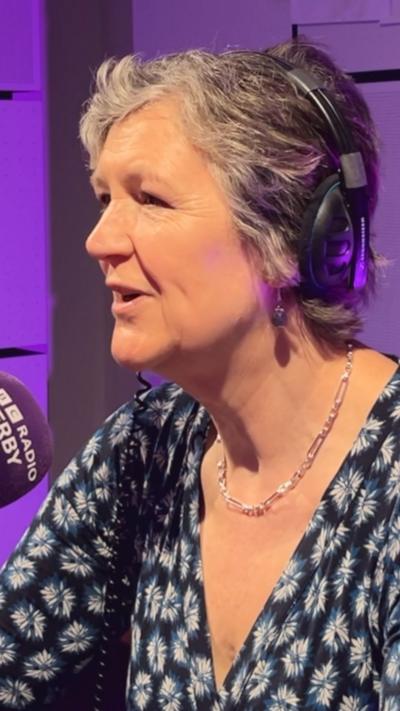 The image shows a person seated in a radio studio environment. The person is wearing over‑ear broadcast headphones and speaking into a microphone that has a BBC Radio Derby mic cover visible on it. The lighting in the studio is a mix of purple and neutral tones, creating a softly illuminated backdrop with acoustic panels behind the individual. The person is dressed in a patterned garment with a dark base color and lighter star‑ or flower‑like shapes. A short, light‑colored necklace and small earrings are also visible. The individual’s hair is short and appears naturally textured with multiple shades visible under the studio lighting.