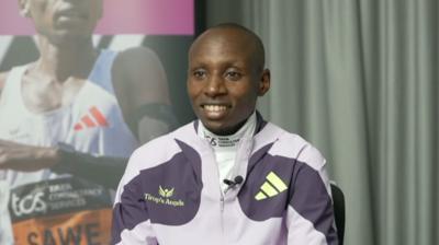 Sabastian Sawe sitting during an interview, wearing a lavender-coloured training top with a curtain behind him