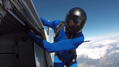 A man wearing a helmet and blue suit clings to the side of a plane.