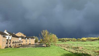 Very dark grey cloudy skies sit above highly contrasted bright green fields and trees with a row of light brick houses to the left