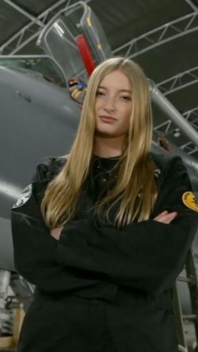 Girl with blonde hair with arms crossed wearing a black flight suit is captured next to a restored grey and red fighter jet