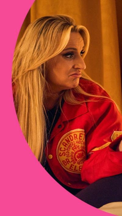 A woman with long blonde hair and a red jacket sits on a bed in a low lit hotel room