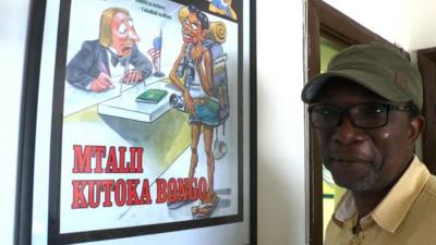 Tanzanian cartoonist James Gayo standing in front of the framed cartoon