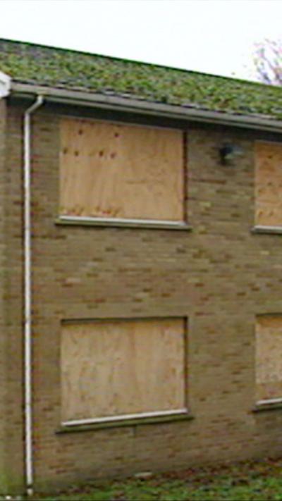 A collection of building boarded up with a blue door in the distance and grass between them.