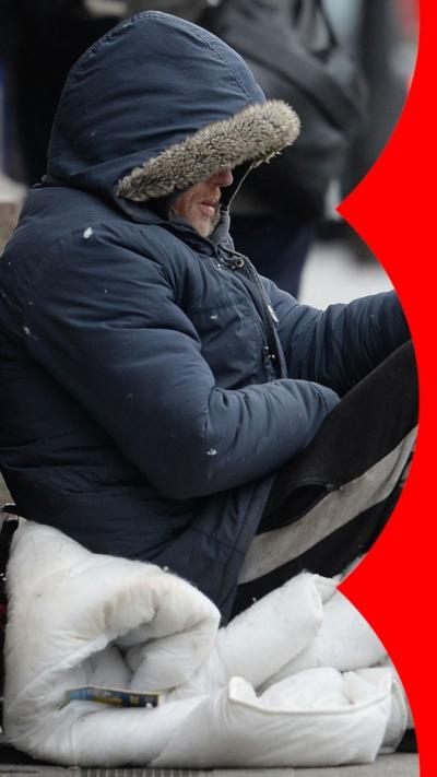 A homeless man wearing a hood holds out a paper cup.