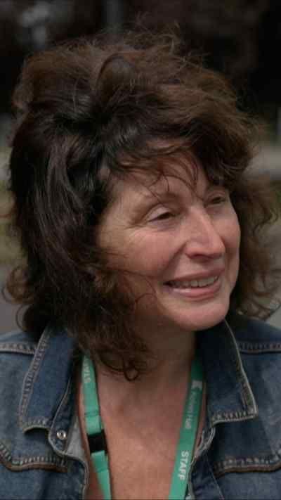 A woman with medium-length curly hair wearing a denim jacket and a green lanyard stands outdoors on a quiet street with trees and houses blurred in the background.