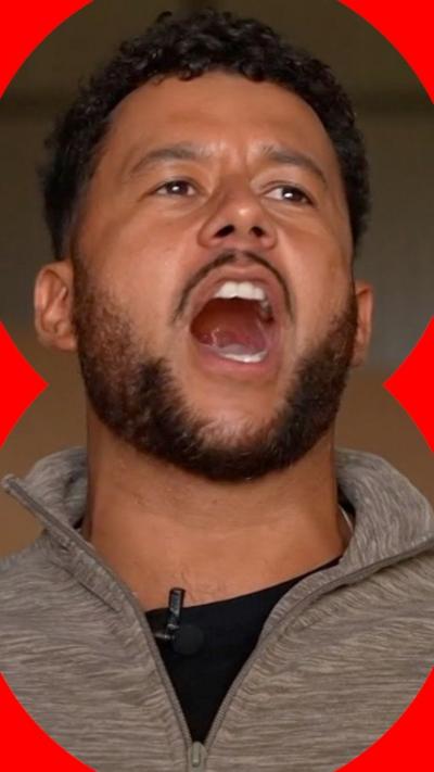 A picture of a man with dark hair singing. He is wearing a beige quarter-zip and a black t-shirt.