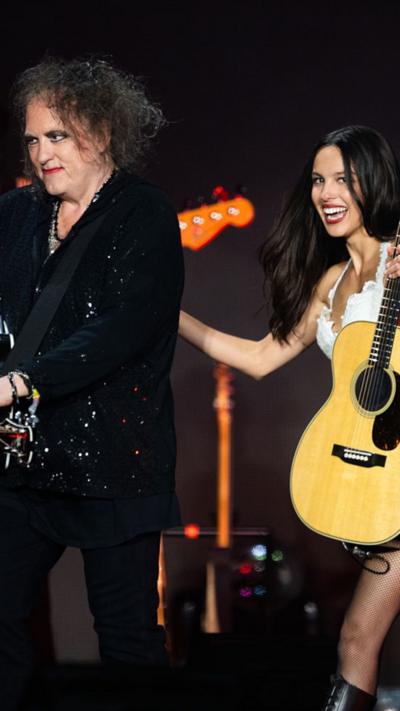 Olivia Rodrigo smiles and holds a guitar as she walks behind The Cure's  Robert Smith at Glastonbury 2025