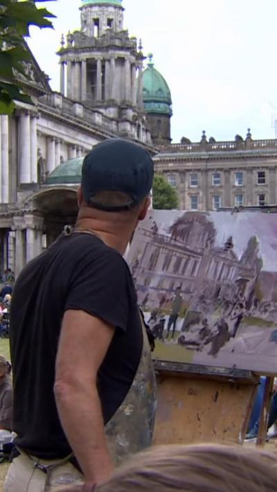 Ed Reynolds is wearing a black t shirt, grey apron and black cap. He is stood in front of a canvas overlooking Belfast City Hall. 