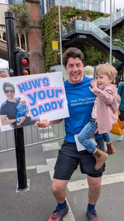 A man in athletic clothes holds a small child while posing on a busy street during the Great Manchester Run, surrounded by crowds and barriers. The man carries a sign reading “HOW’S YOUR DADDY” with a photo, with a brick bridge, greenery-covered wall, and a staircase with onlookers in the background.