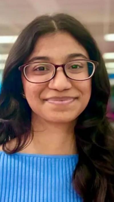 A woman wearing a blue top stands and smiles at the camera. She wears glasses and has long dark hair.