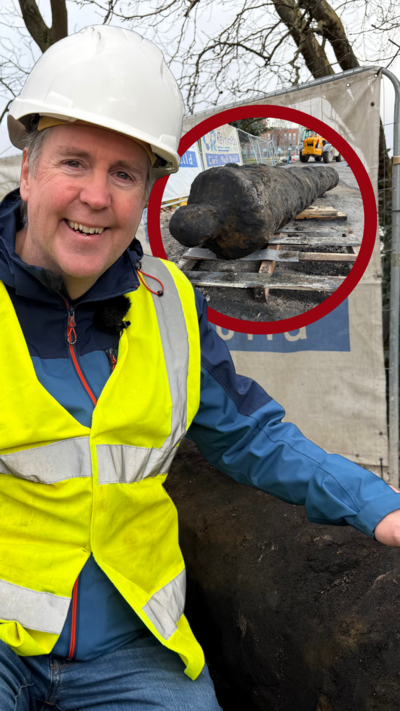 A man wearing a white hard hat and yellow hish-vis jacket is kneeling beside a cannon that has been dug up from the ground.