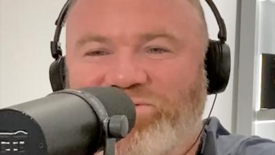 A man, Wayne Rooney, has a beard and is wearing headphones and talking into a microphone