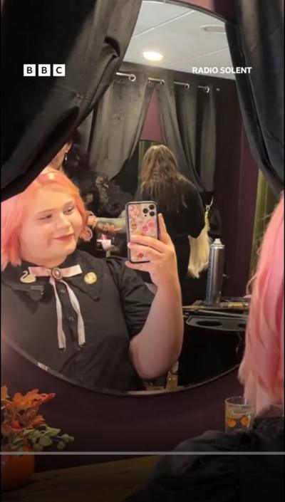 Smiling woman with pink hair taking a selfie in a mirror flanked by black curtains