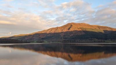 A Cumbrian hill, half in sunlight and half shadow, reflected in water