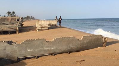 Two people walk along a beach in Ghana where houses are being buried in sand