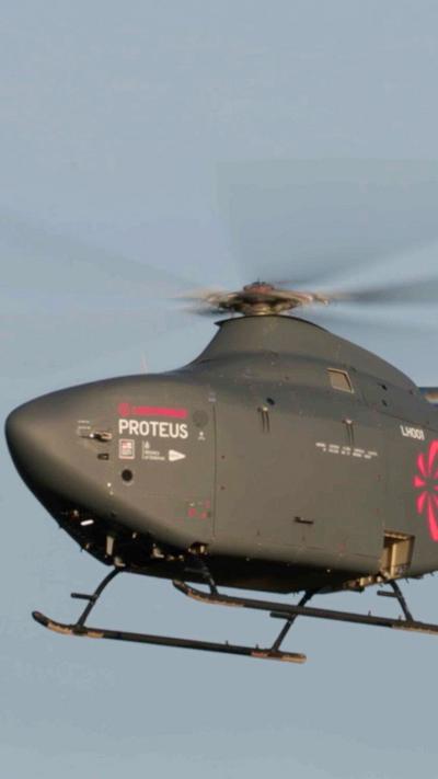A helicopter with no windows flies, with its rotors a blur. It is a dark grey-green and has red markins on it.