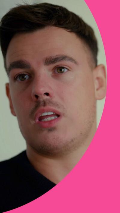 A man with short dark hair a black tshirt looks at someone off camera with a serious expression