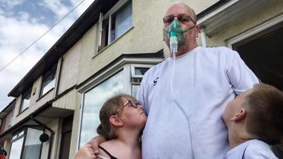 Rob Hill wearing an oxygen mask, embracing two of his children