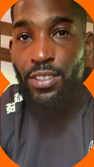 A man with short black hair and a short beard looks at the camera, he wears a black top