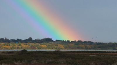 There are brown fields and a river in the fore and mid-ground but above a tree-filled horizon a huge arc of a rainbow rises up into the sky.