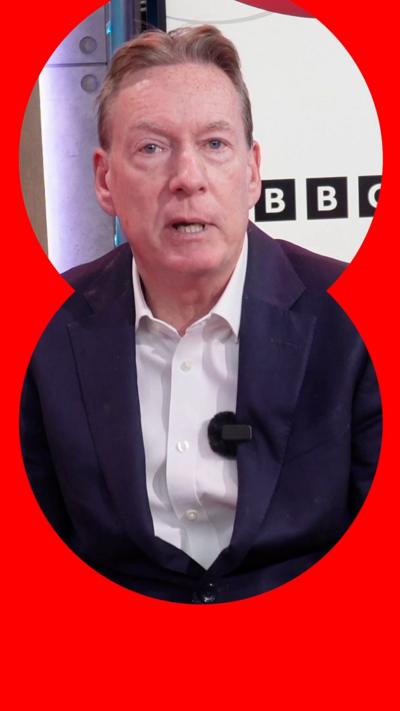 Frank Gardner is sat down and wearing a navy suit with a white shirt. Behind him is a screen with the BBC logo on.