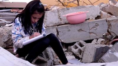 Girl writes in book as she sits among the rubble