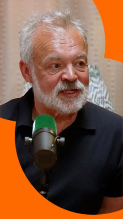 Graham Norton wearing a black polo shirt. He's sat in front of a green mic.