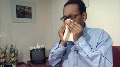 Lizo Mzimba blowing his noise, sitting on a sofa.  Small television set and flowers sit in the background, with a couple of picture frames on the wall.