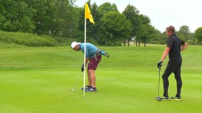 Man stands on golf green watching another man taking a short shot into the hole on a sunny day.