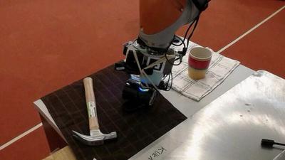 A robotic arm on a table, on the left-hand side is a hammer, on the right-hand side is a mug on top of a tea towel