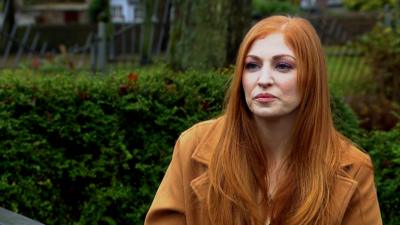 A red-haired woman - Dawn Fernand - sits in front of a green hedge wearing an amber-coloured coat and lilac eyeshadow.