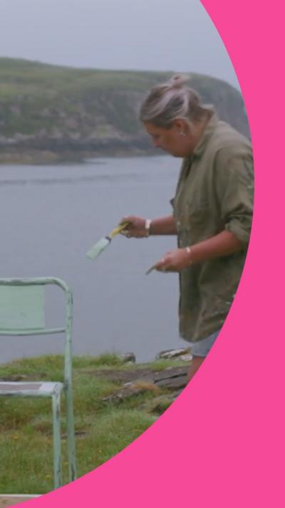 A woman paints a chair outdoors, with the sea behind her