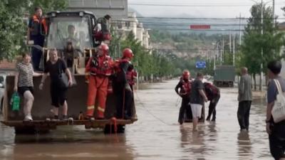 People stand on an excavator vehicle's front loader bucket in the middle of a flooded road.