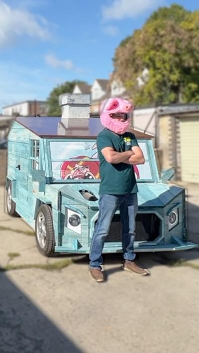 A man in a pink motorbike helmet stands in front of his shed car.