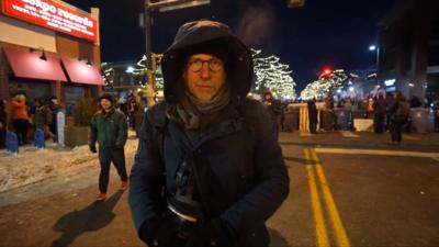 Tom Bateman wearing a hooded coat near protests on a blocked off street in Minneapolis