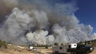 Plumes of thick smoke rise into the air as wildfires rage in California. A truck and trailer drive along a desert road in the foreground.