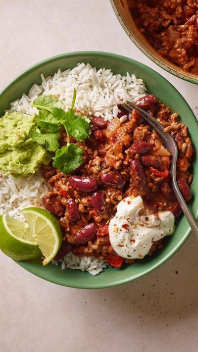 A white bowl containing rice, tofu chilli, guacamole, lime and tortilla chips.